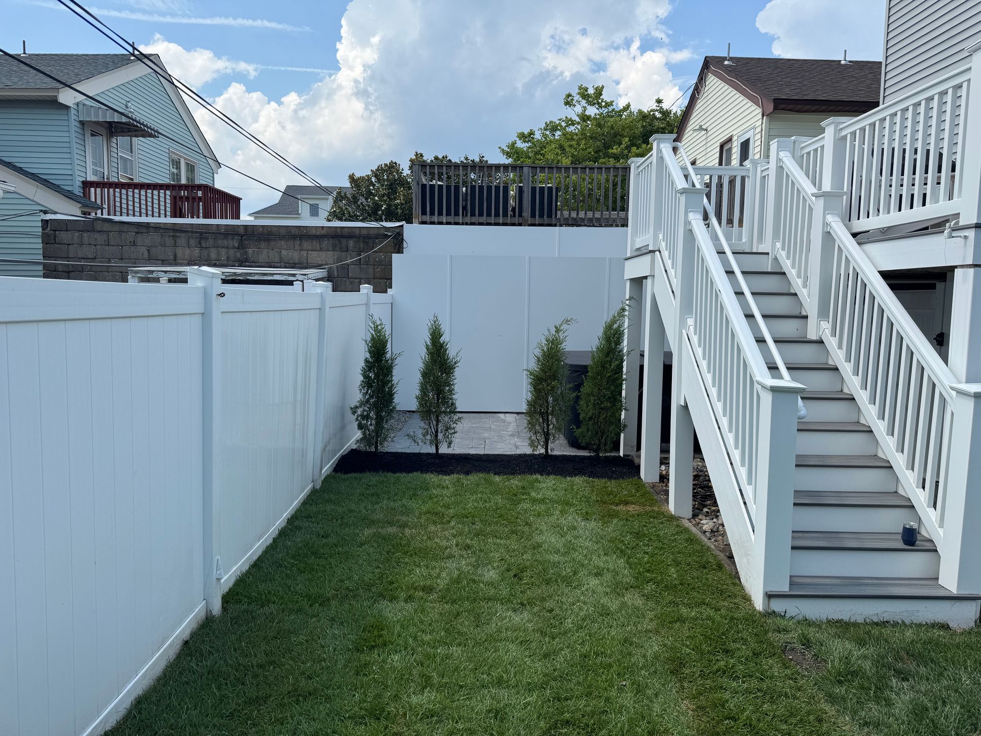 White fenced backyard with stairs and small evergreen trees along a white wall. Green grass.