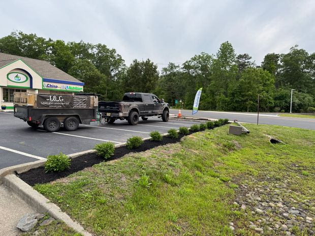Dark truck with trailer parked outside a store, boxes on trailer, bushes in front.