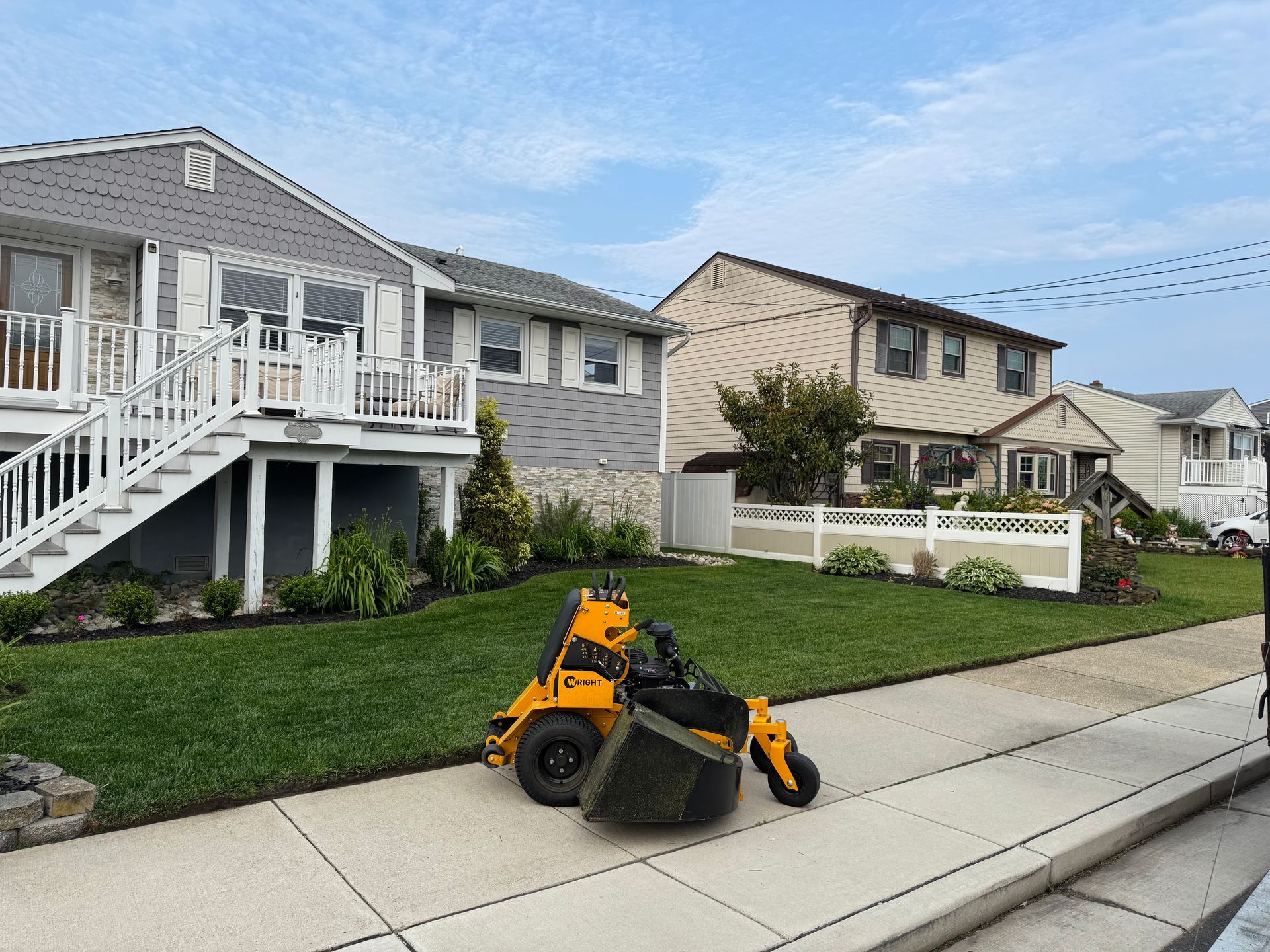 Yellow robotic lawnmower on a sidewalk, cutting grass in front of houses on a sunny day.
