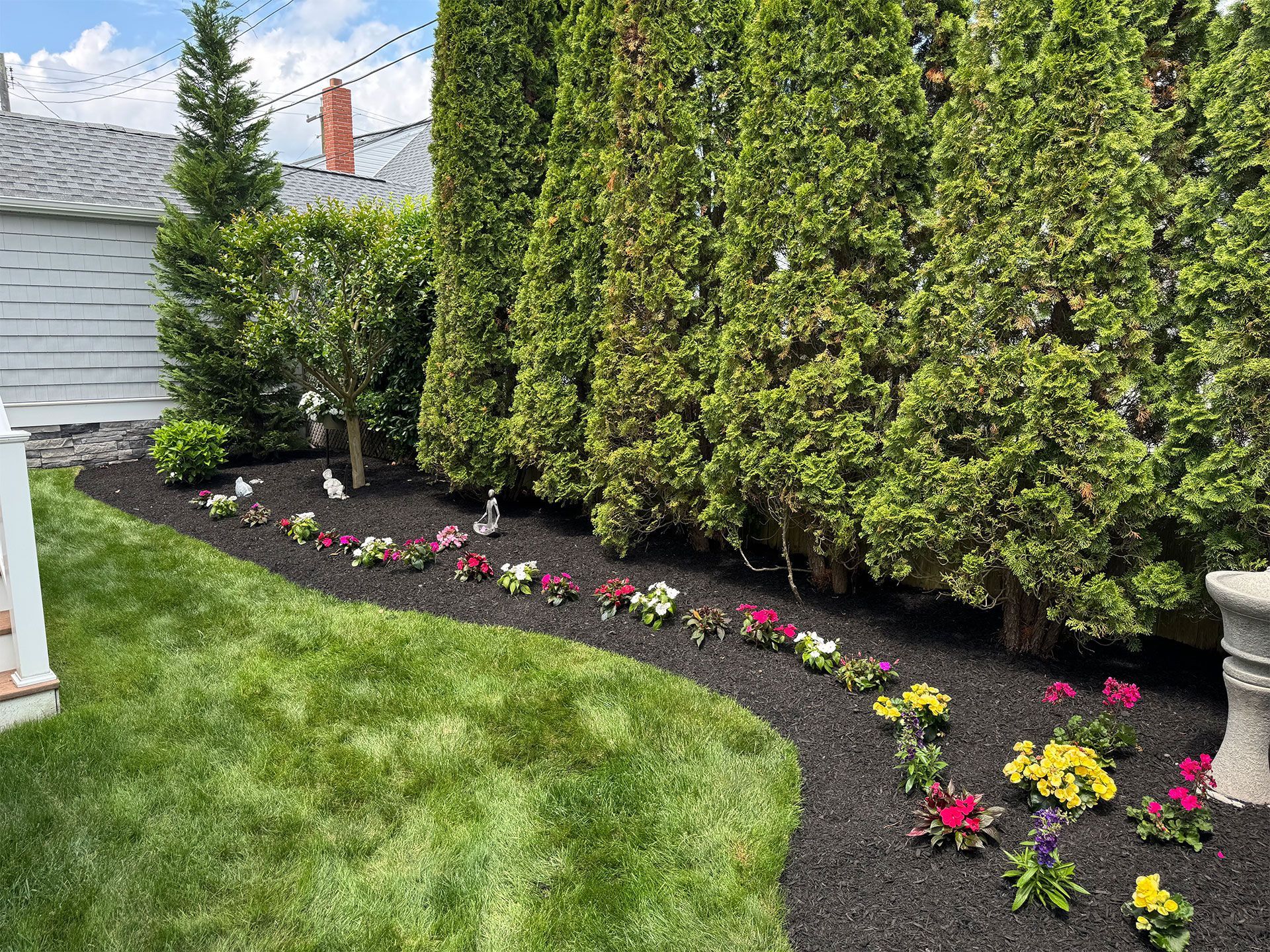A garden bed with colorful flowers and dark mulch borders a green lawn, next to tall evergreen trees.