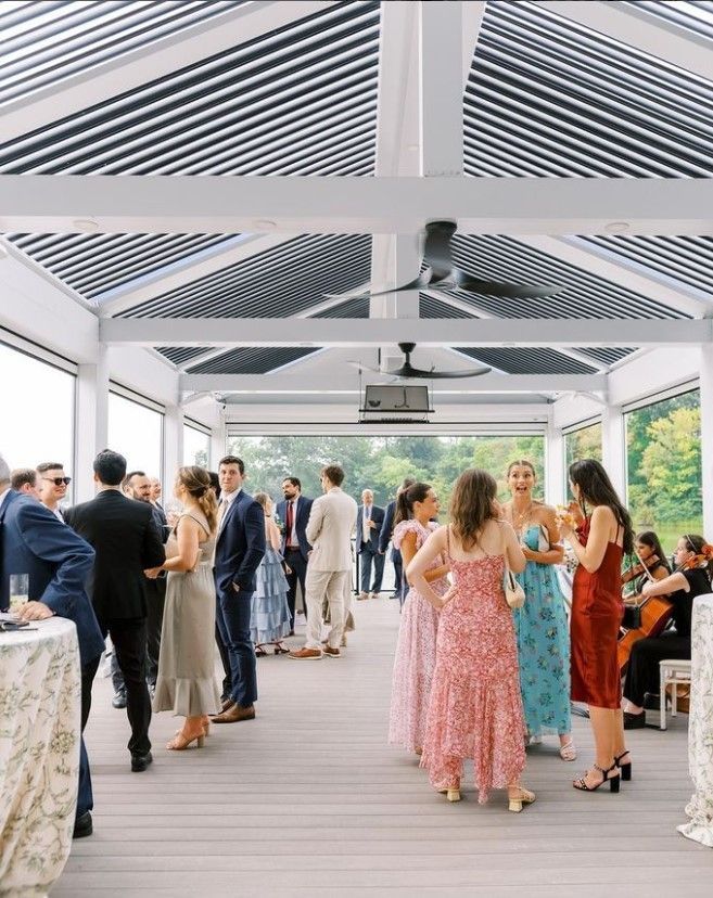 Guests mingle in a bright covered patio at a formal event, with women in colorful dresses and men in suits.