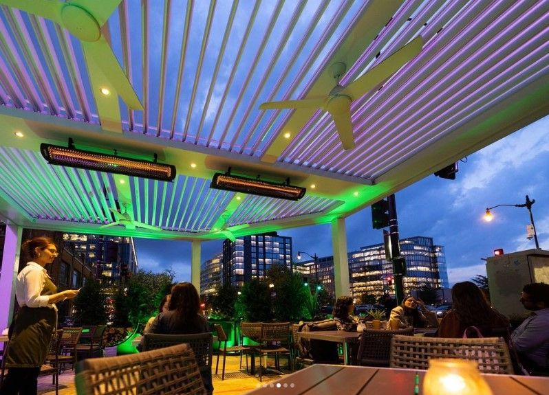 Outdoor rooftop bar at dusk with neon-lit ceiling, city skyline, and seated patrons at tables