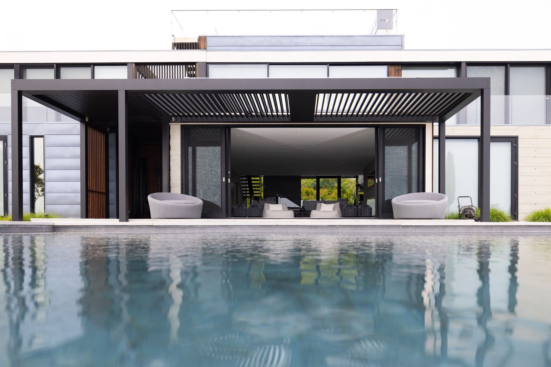 A modern home with a dark metal pergola over a patio, overlooking a calm swimming pool.