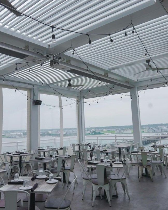 Open-air rooftop restaurant with white tables and chairs overlooking a waterfront under a slatted roof