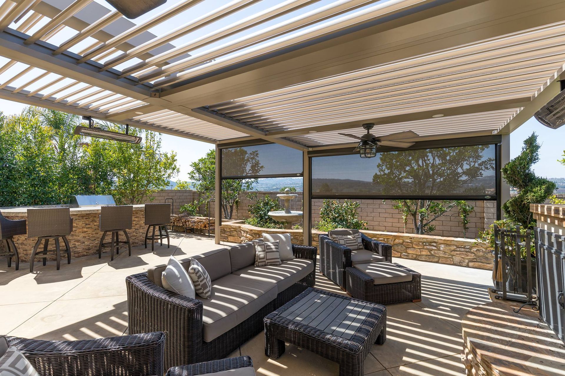 A shaded outdoor patio with a wicker sofa, coffee table, lounge chairs, a bar, and retractable privacy screens.