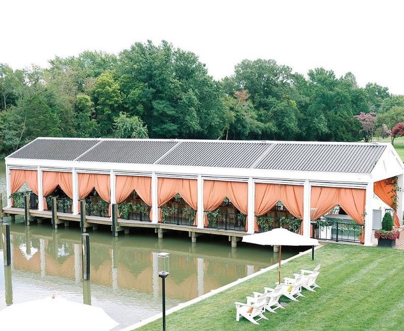 Open-air pavilion with peach drapes beside a pond, surrounded by trees and white lawn chairs