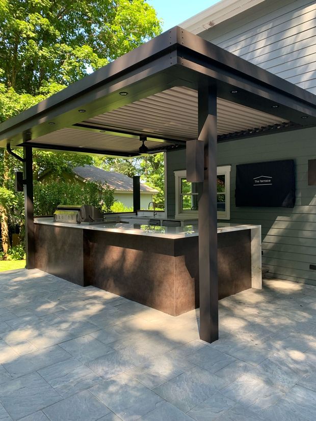 Outdoor kitchen with a stone counter, black metal pergola, and ceiling fan on a stone patio next to a house.