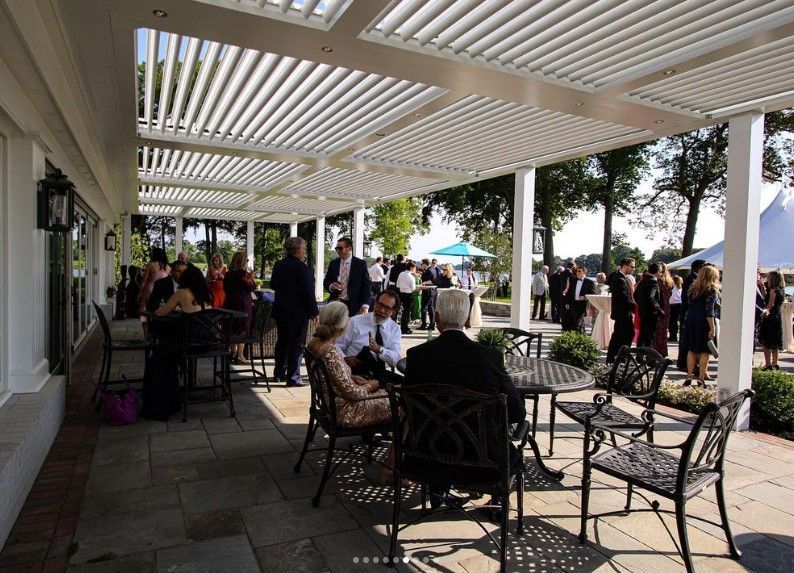 People seated and standing on a patio under a white pergola at an outdoor gathering