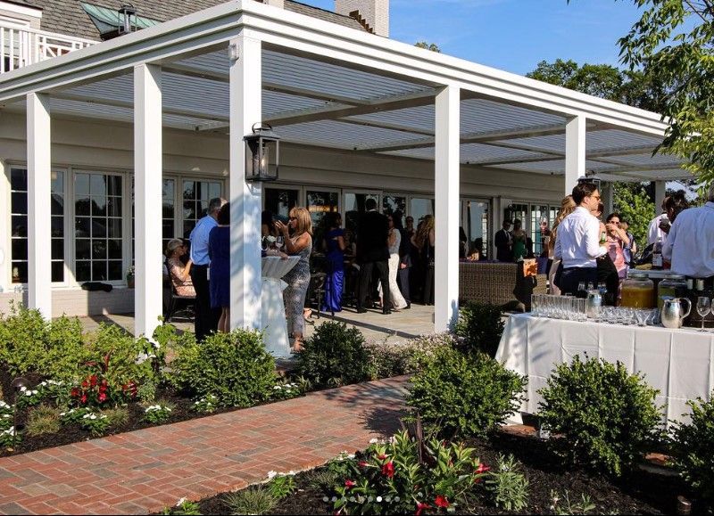 Outdoor wedding reception under a white pergola with guests mingling on a brick patio and garden flowers nearby