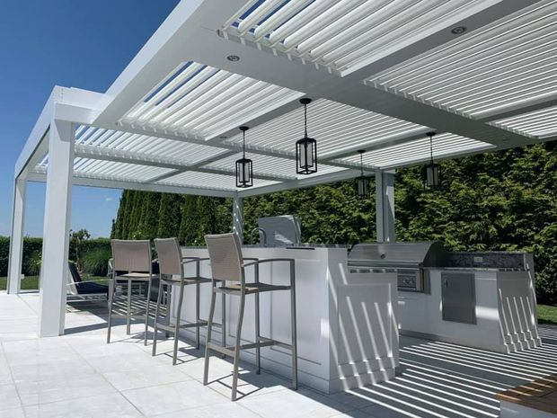 A modern white outdoor kitchen and bar area featuring a louvered pergola roof, hanging lights, and stools on a patio.