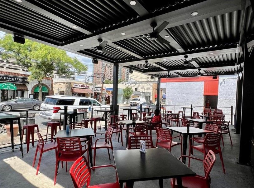 Outdoor restaurant patio with red chairs and black tables under a striped awning, street visible beyond