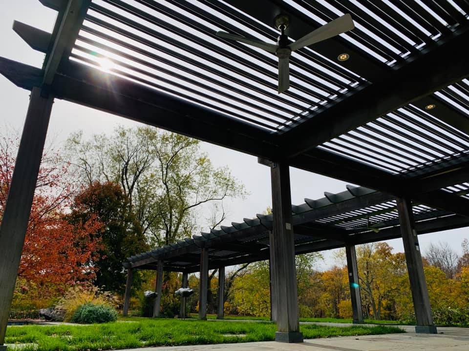 A wooden pergola with a ceiling fan overhead, looking out onto a park with autumn trees and a grassy lawn.