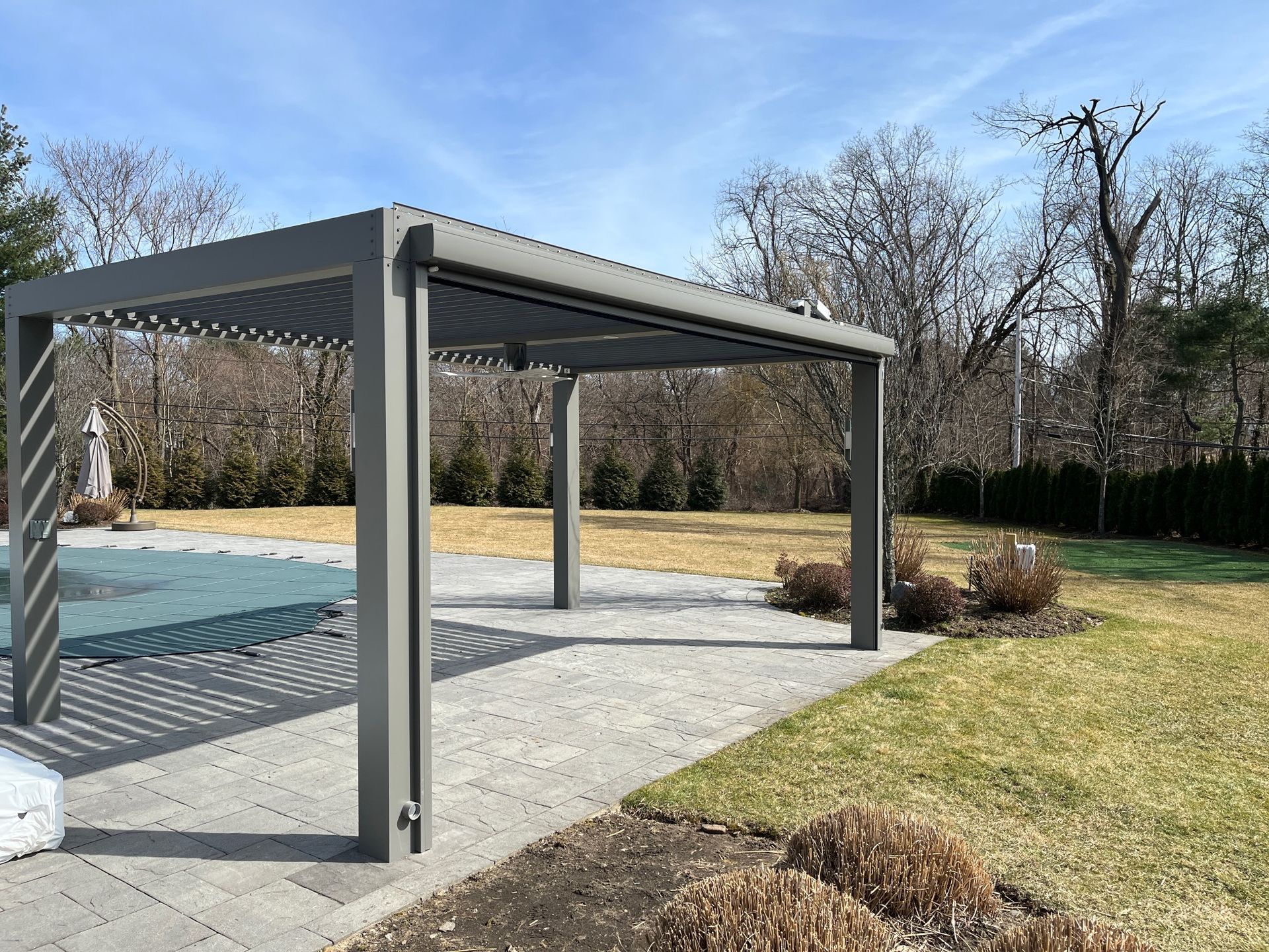 Gray pergola beside an outdoor pool on a paved patio, with grassy yard and leafless trees in the background