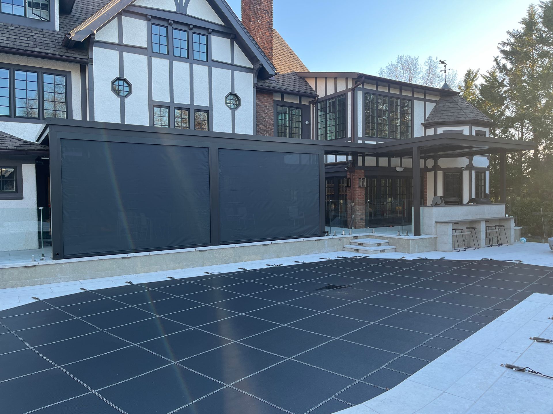 A Tudor-style house with a black-covered driveway or patio in front, framed by snow and winter sunlight.