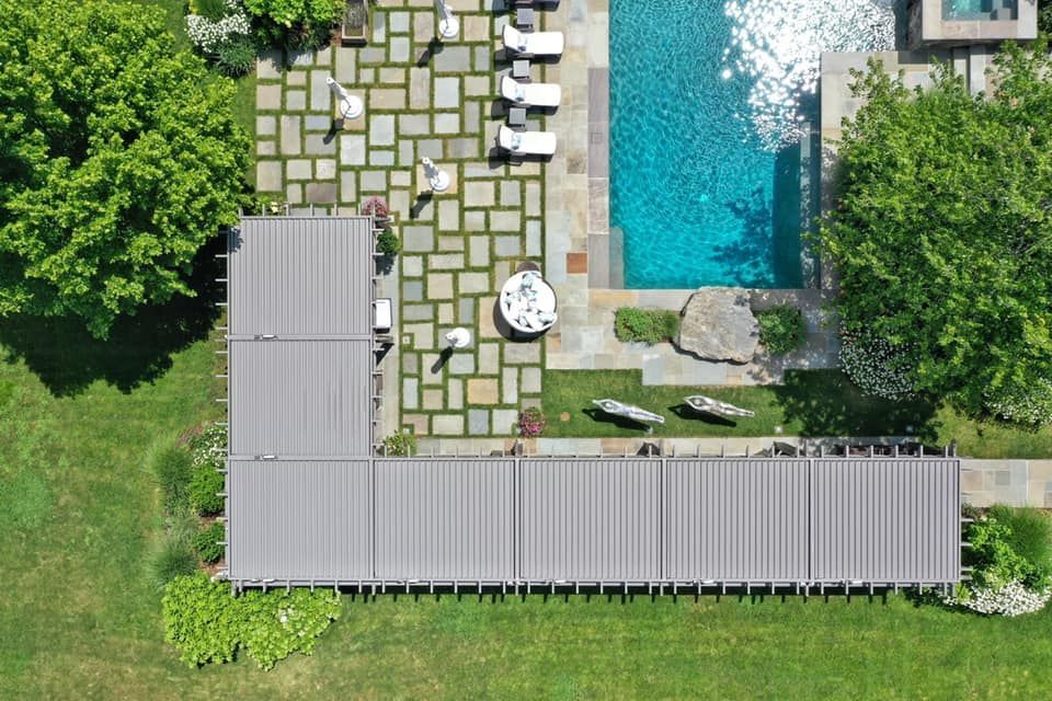Aerial view of a rectangular L-shaped building with a metal roof next to a stone patio, pool, and surrounding green lawn.