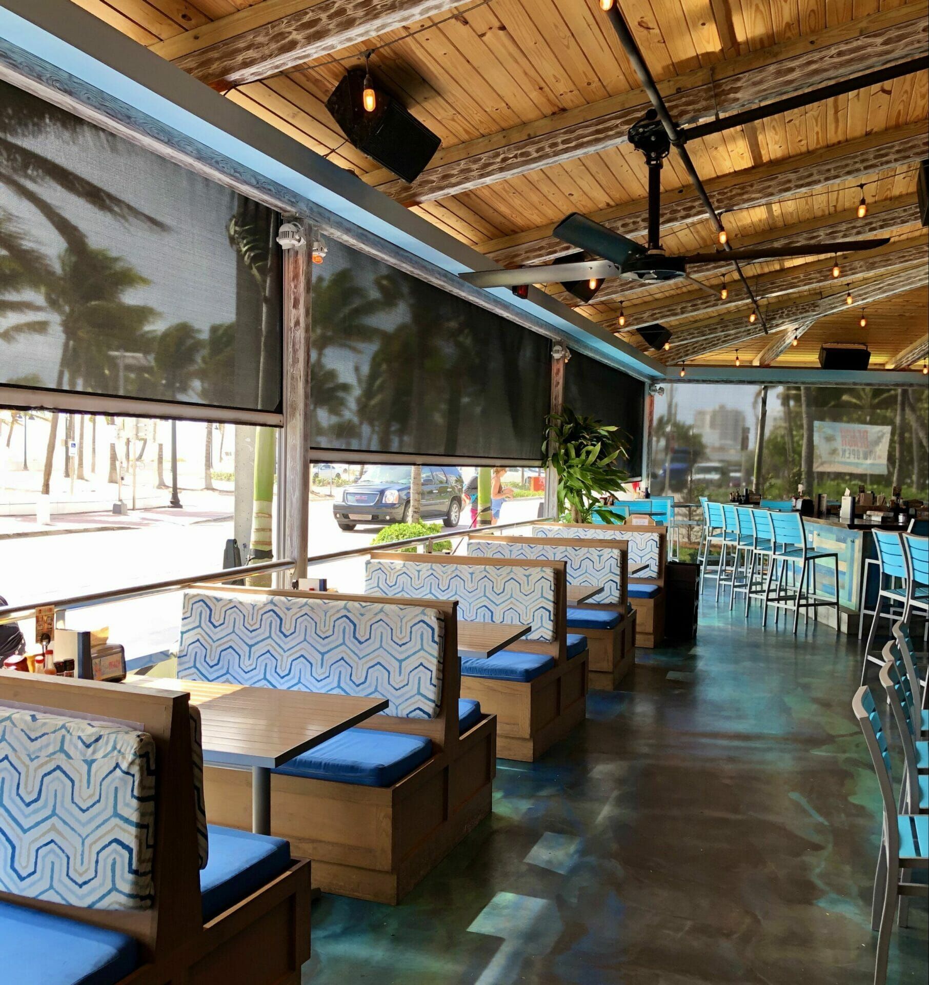 Open-air restaurant with blue cushioned booths, wooden ceiling, and sunlight streaming in from the windows