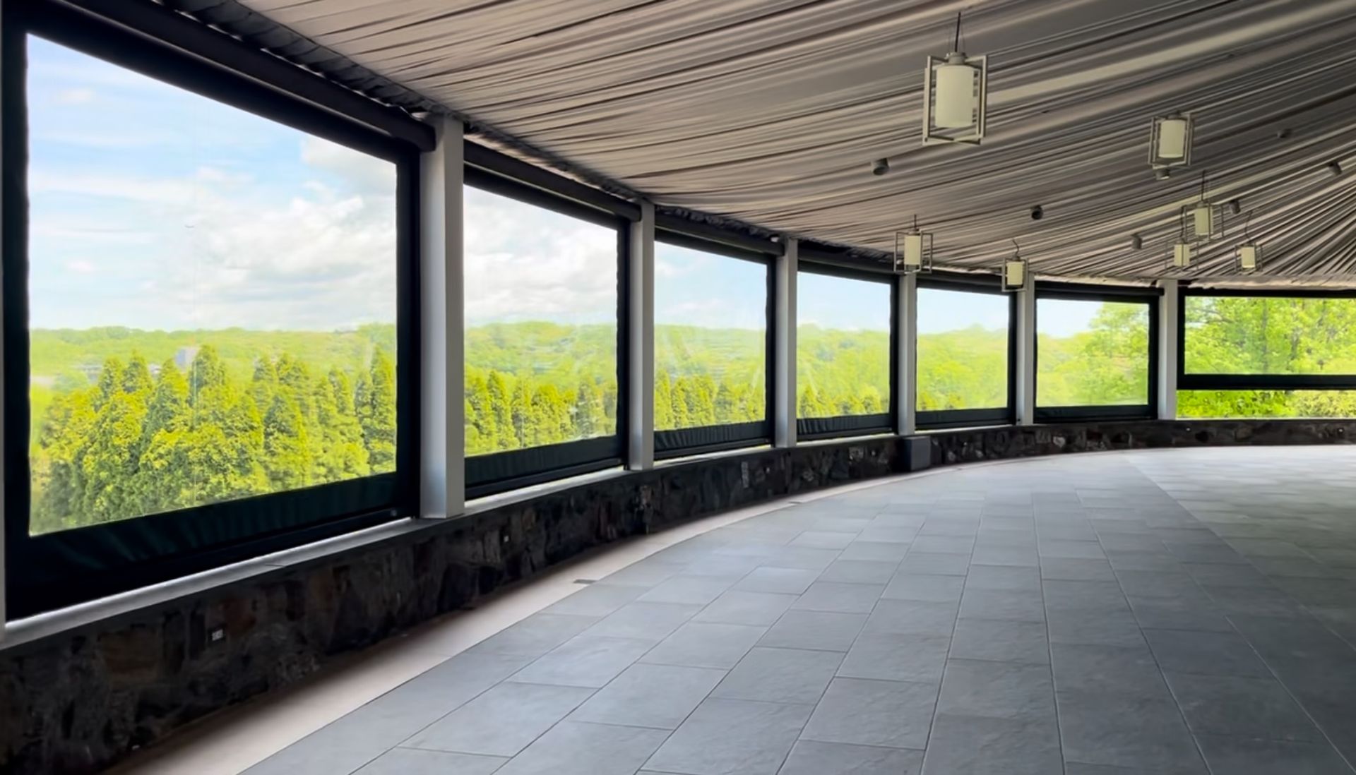 Curved covered terrace with large black-framed windows overlooking green landscape and blue sky
