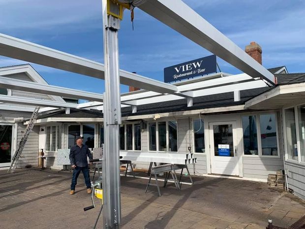 A person stands on a patio under a large, white, partially assembled metal frame structure in front of a building.