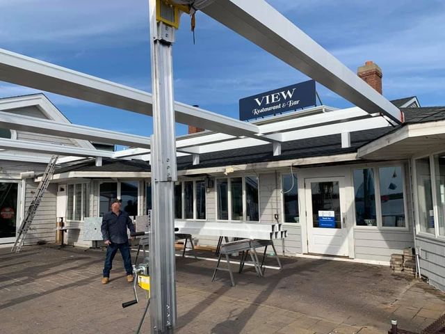 A person stands on a patio under a large, white, partially assembled metal frame structure in front of a building.