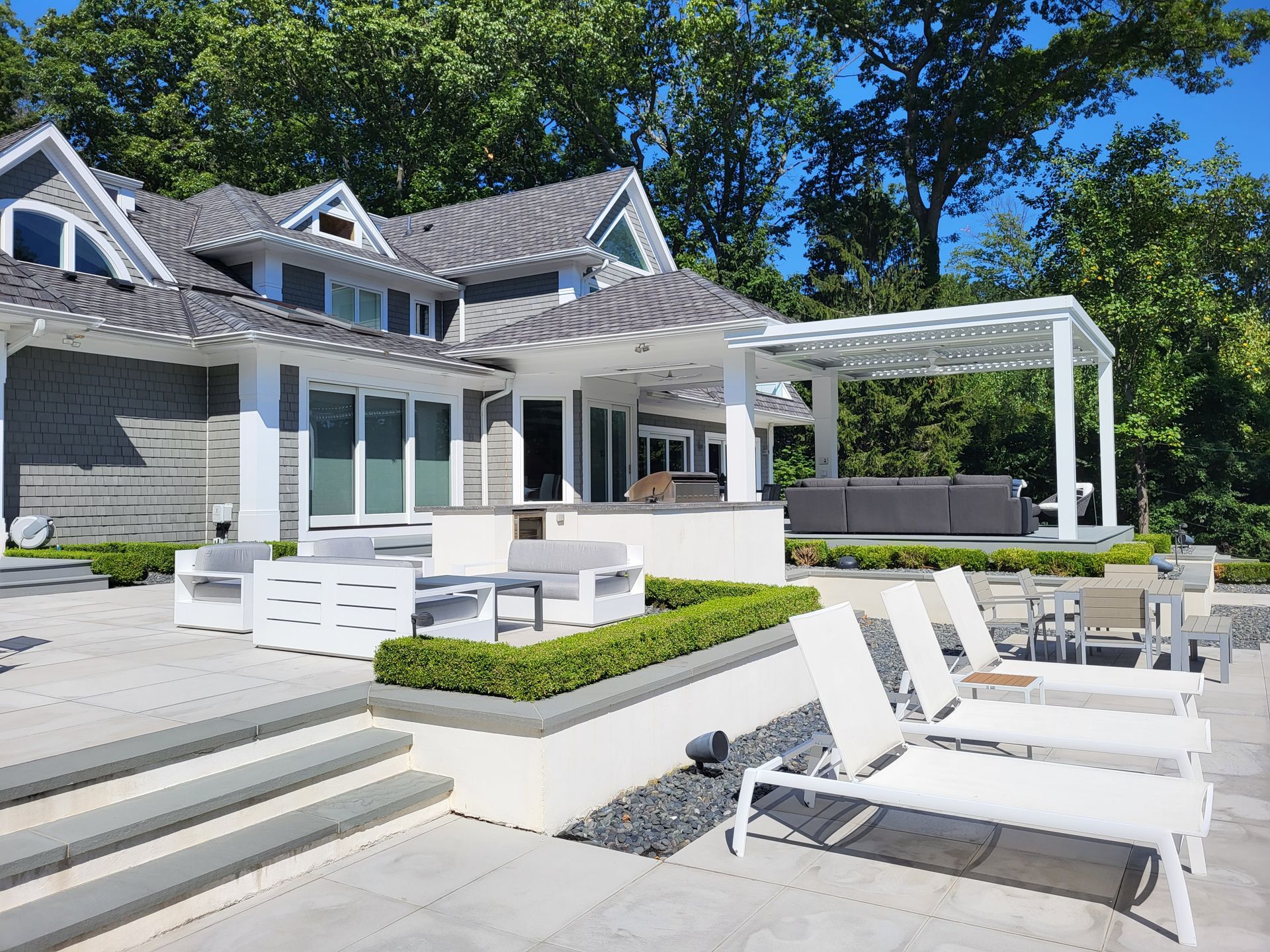 Modern white patio with loungers, pergola, and outdoor seating beside a gray house