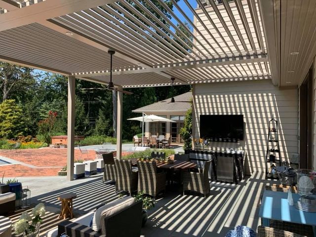 A covered patio with a slatted roof, dining furniture, a wall-mounted TV, and a view of a garden and pool area.