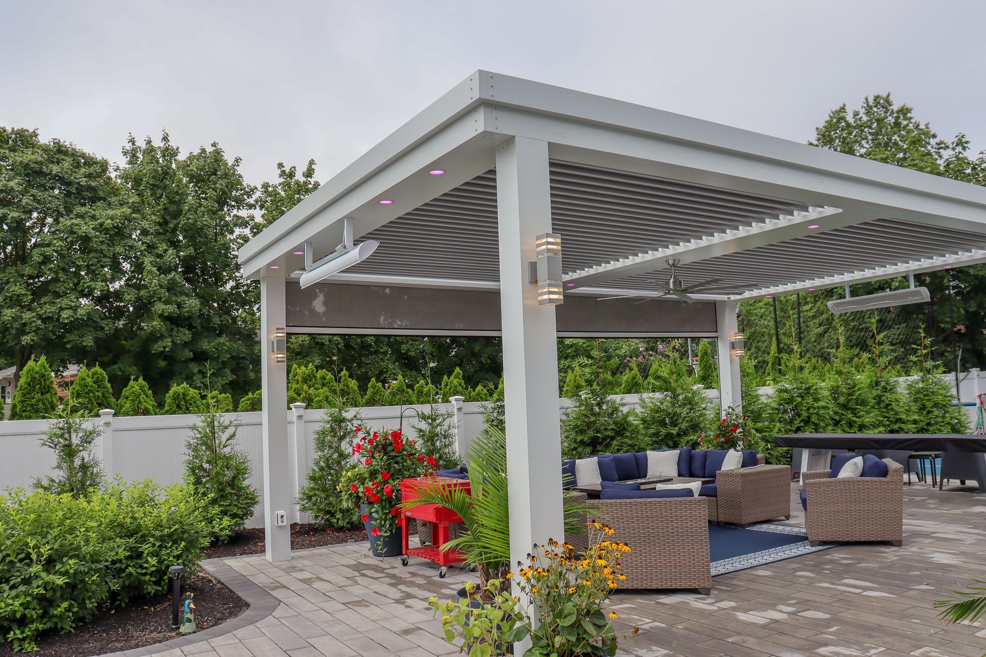 Modern backyard patio with white pergola, wicker seating, and red chairs beside lush greenery