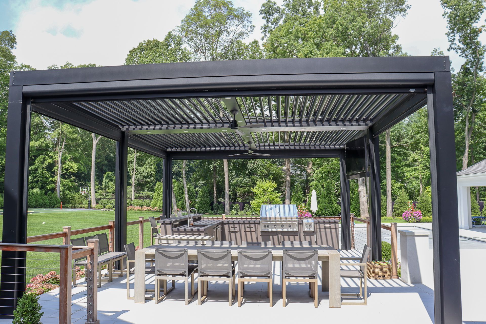 Covered outdoor seating area with tables and chairs under a black pergola in a park-like setting