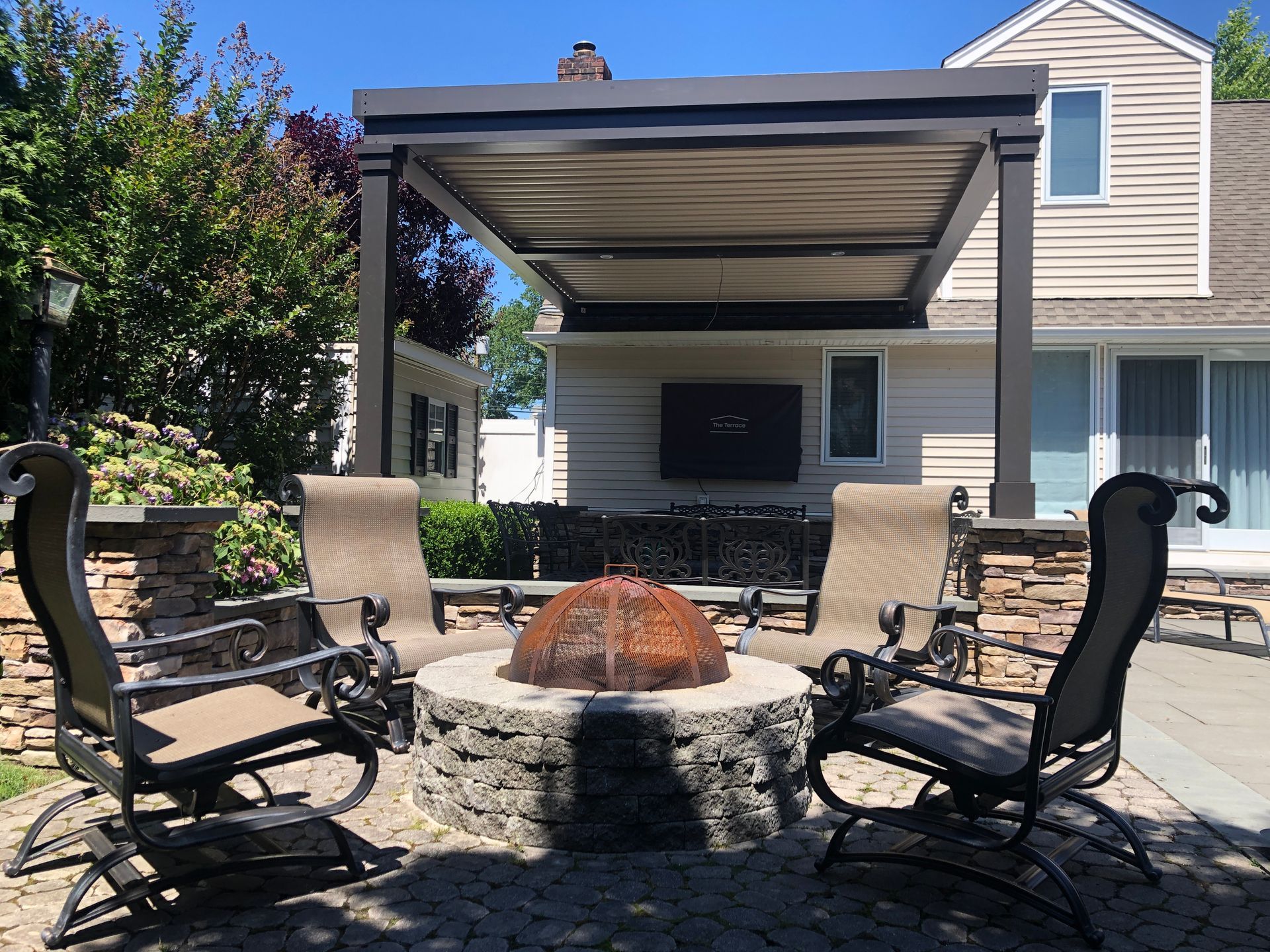 Backyard patio with a fire pit, two cushioned chairs, and a covered pergola beside a house.