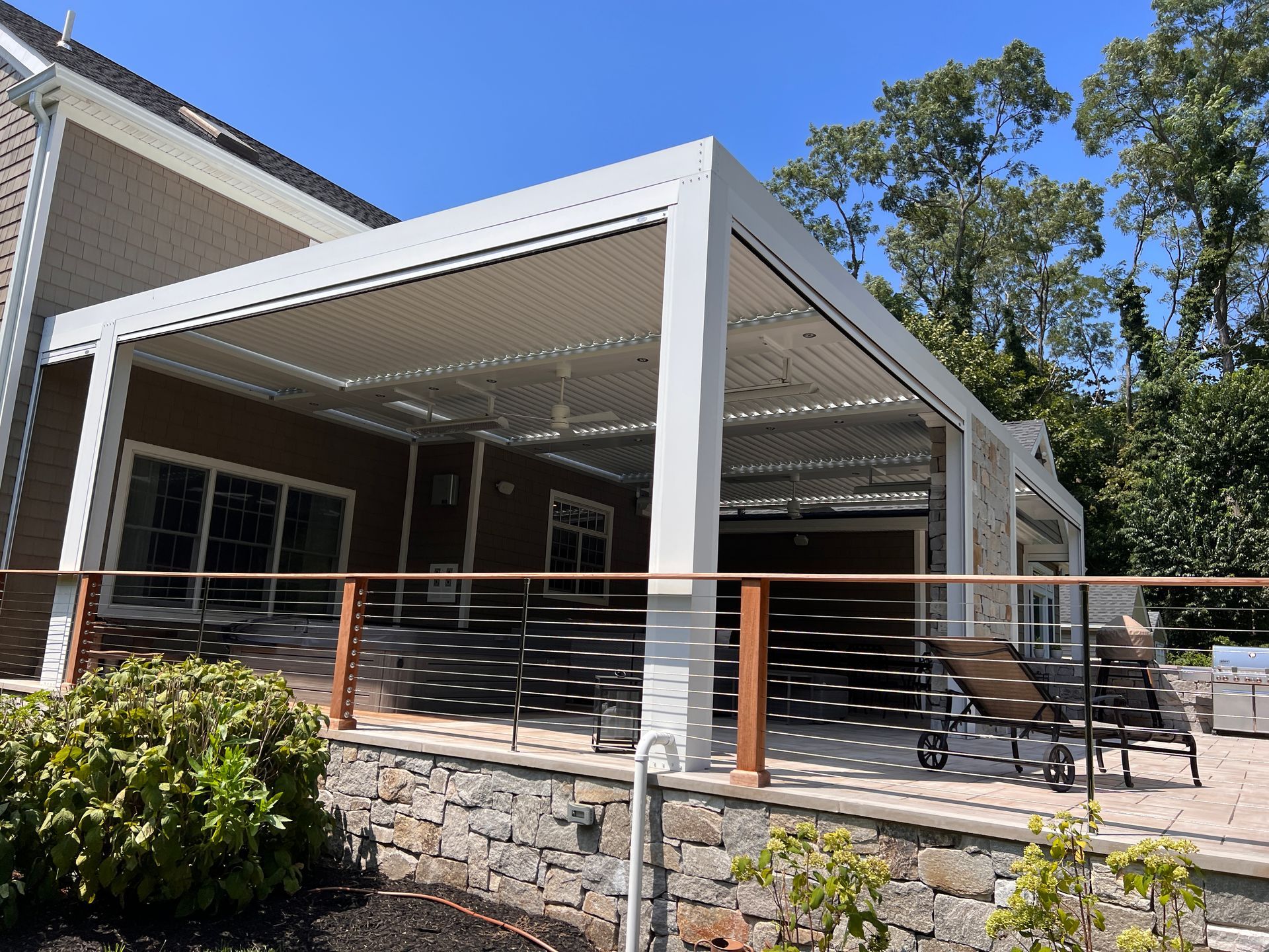 Covered outdoor patio with white beams, stone base, and metal railing beside a house on a sunny day