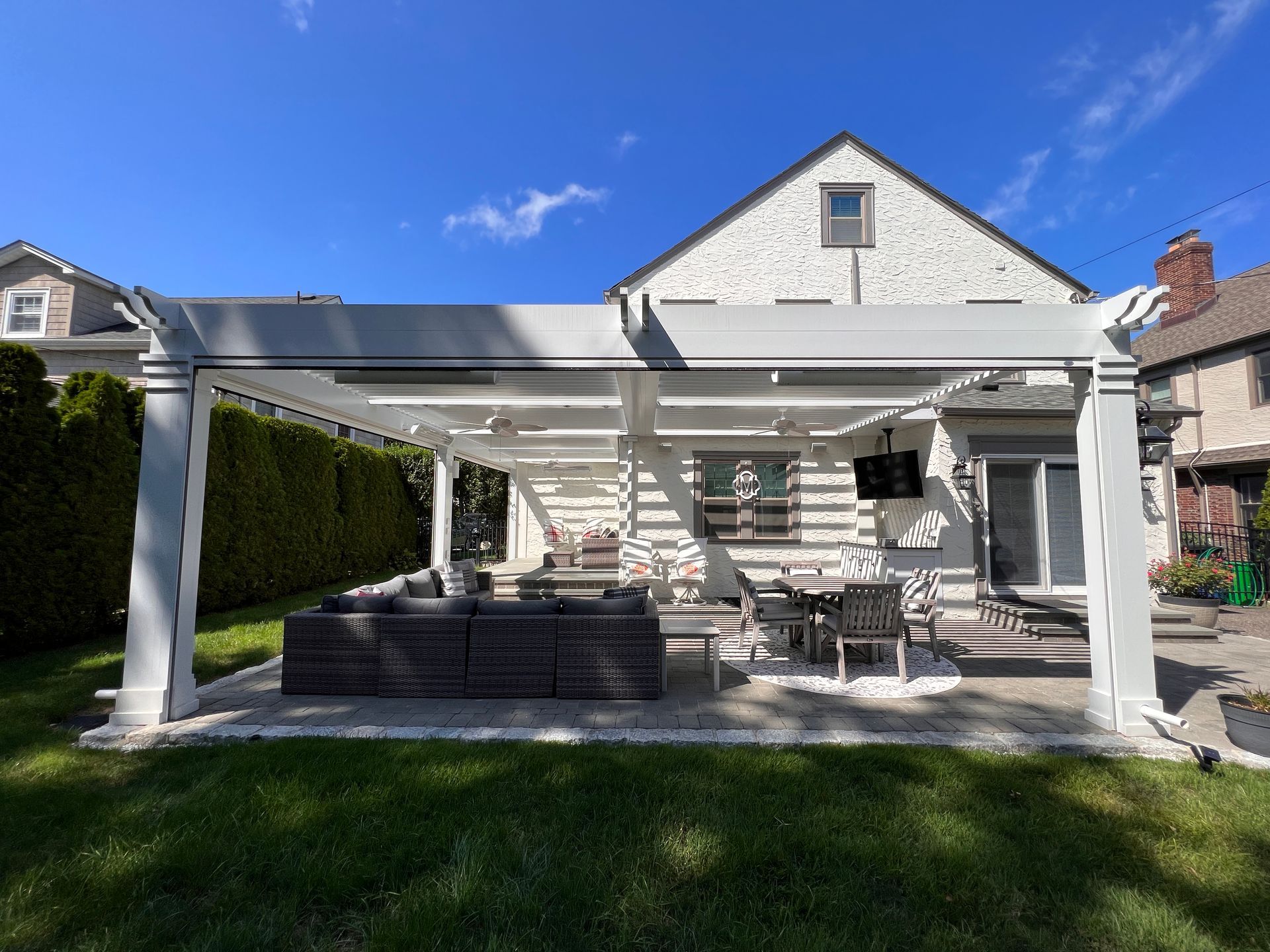 Backyard patio with white pergola, outdoor seating, grill, and green lawn under a blue sky