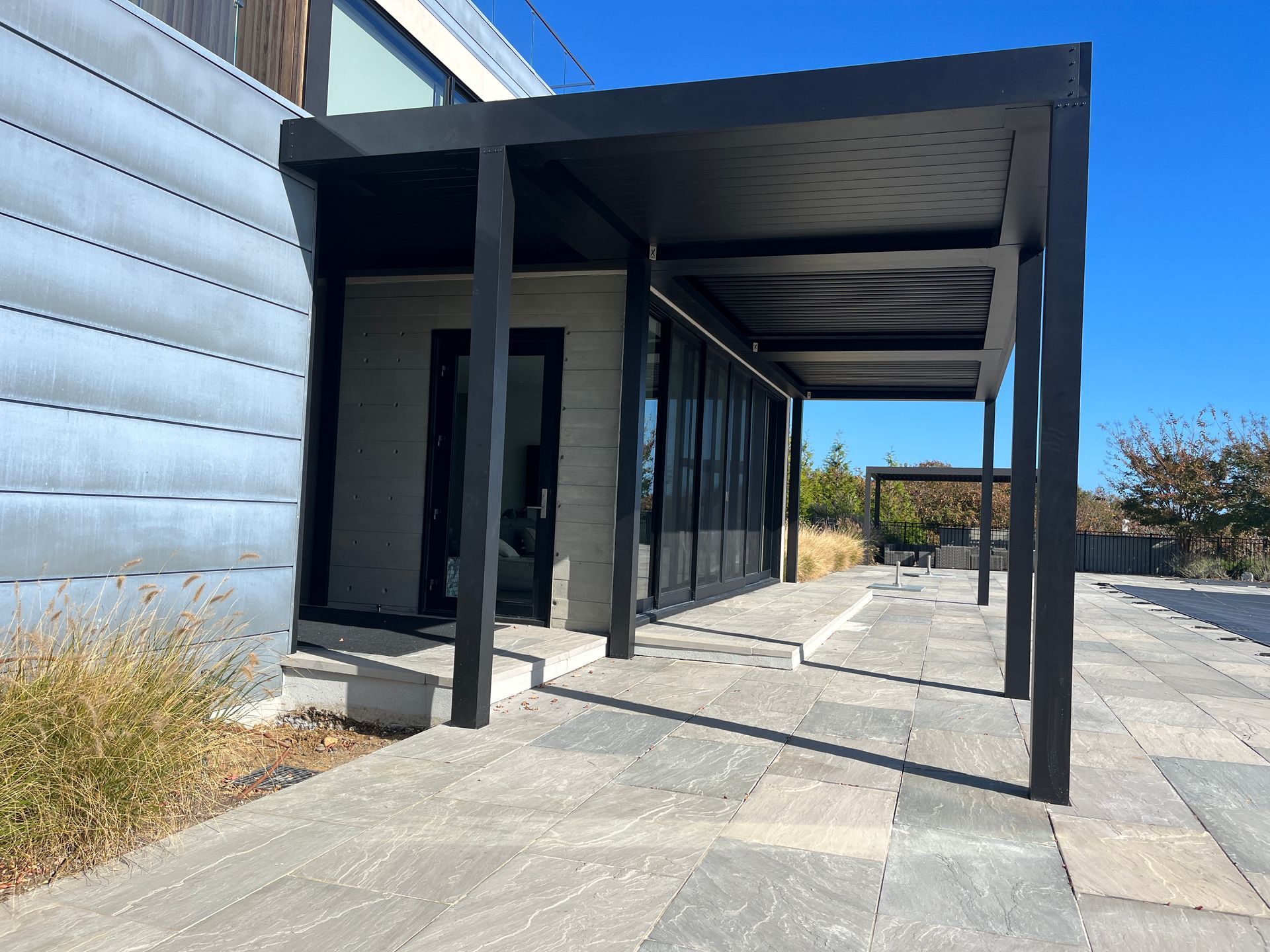 Modern black-framed building with a covered walkway and concrete patio under a clear blue sky