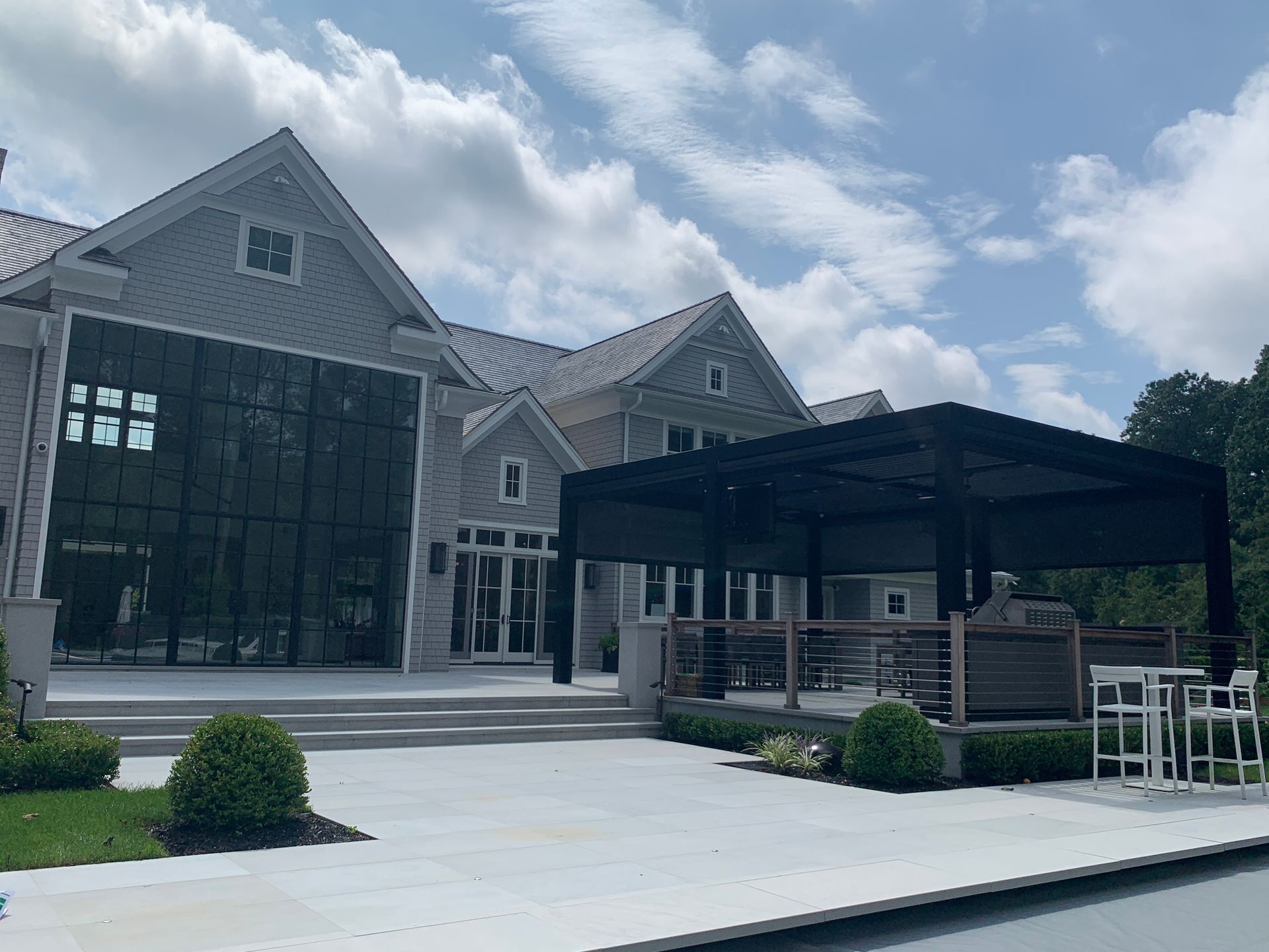 Modern stone building with large windows, covered patio, and paved courtyard under a partly cloudy sky