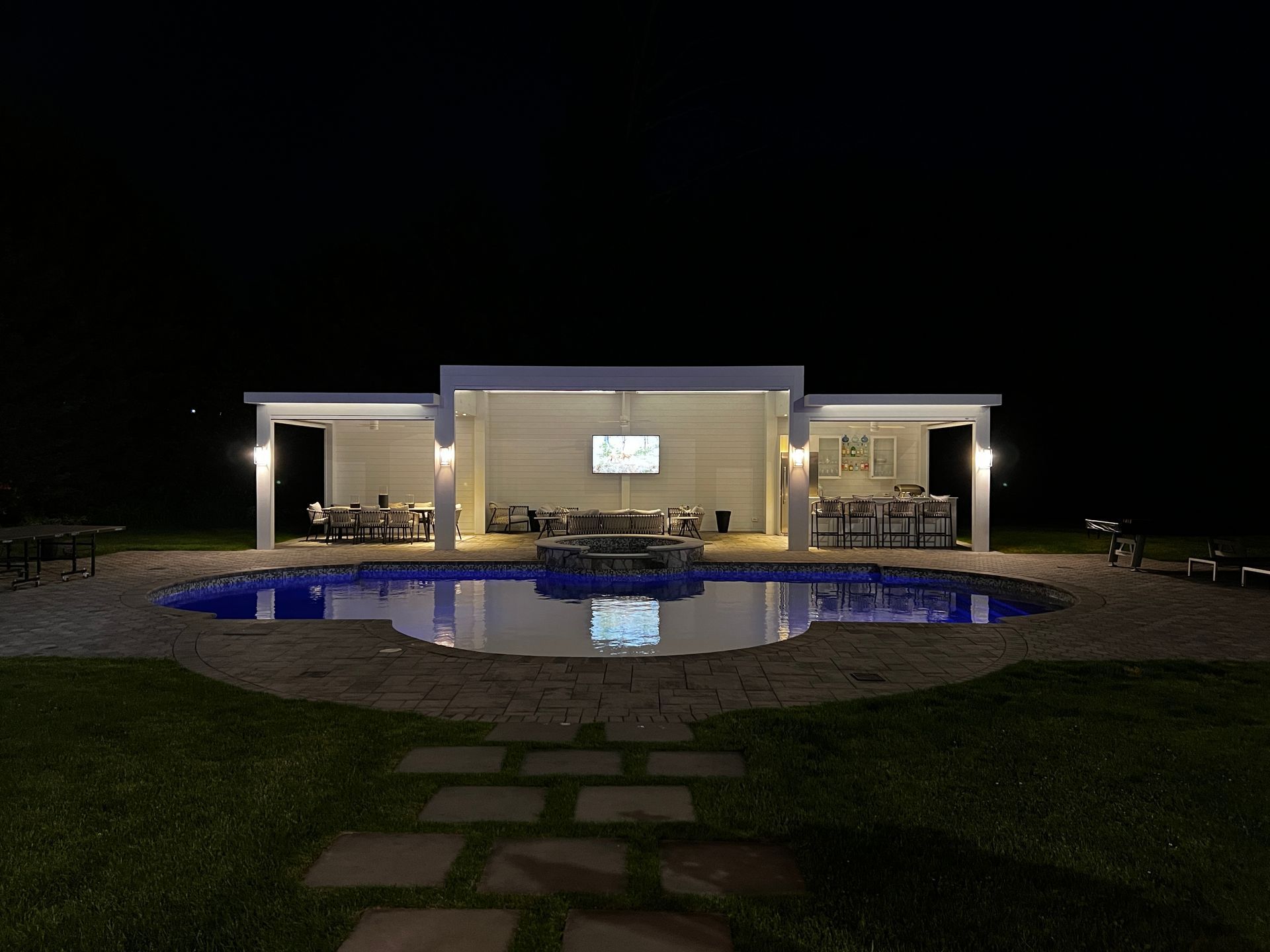 Nighttime view of a modern poolside home with lit patio and stone path leading to the water