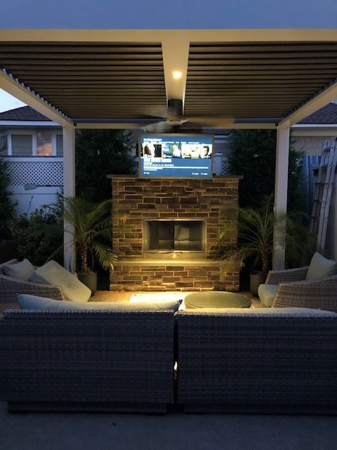Covered outdoor patio with brick fireplace, cushioned seating, and a wall-mounted TV at dusk