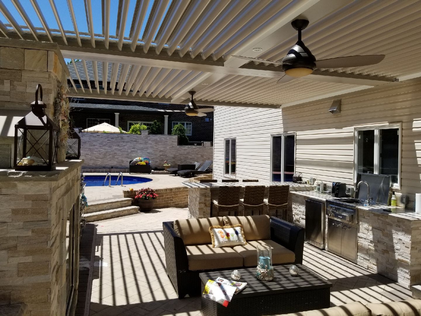 Covered patio with wicker seating, grill, ceiling fan, and striped shadows overlooking a pool.