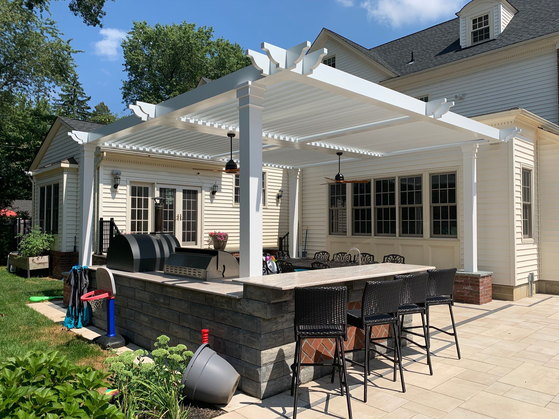 White pergola-covered patio with outdoor dining, grill, and stone bar beside a house
