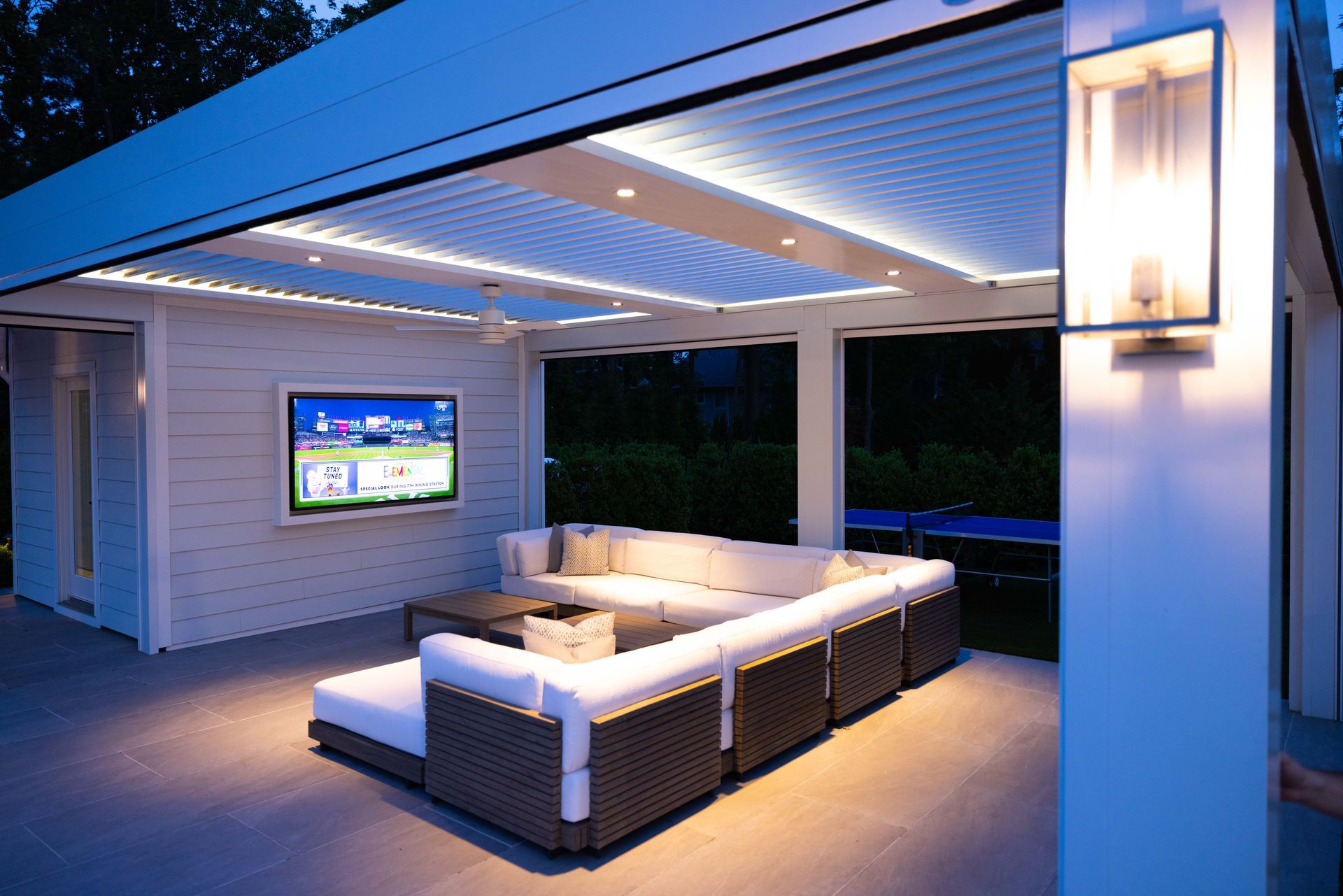Modern patio with sectional sofa, wall TV, and blue evening lighting under a covered pergola