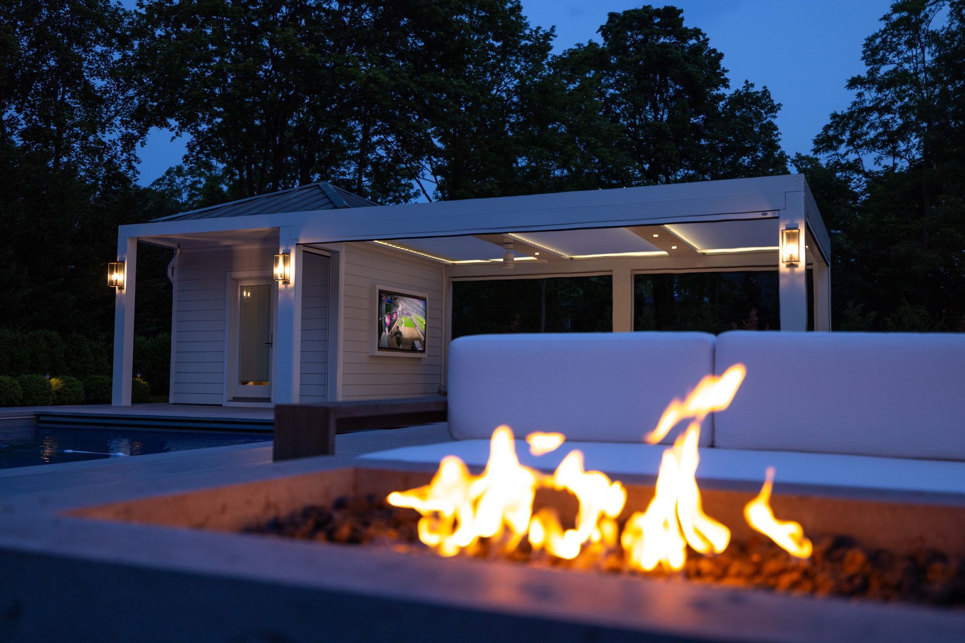Modern patio with fire pit in front of a white outdoor seating area at dusk