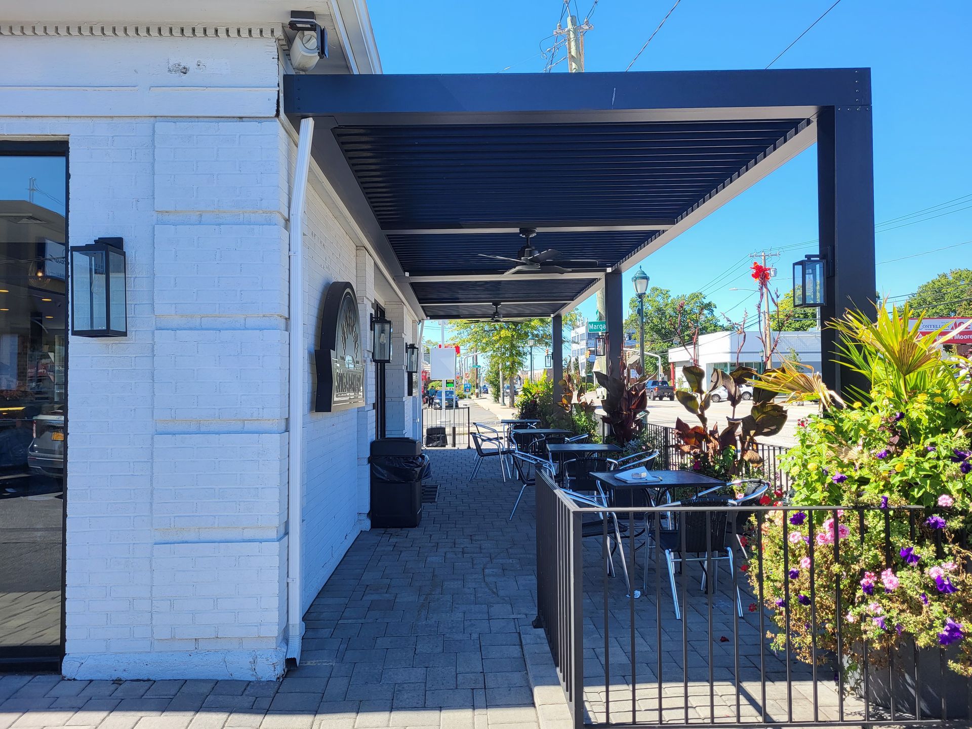 Blue awning over a sidewalk patio with tables, chairs, and potted flowers beside a white building