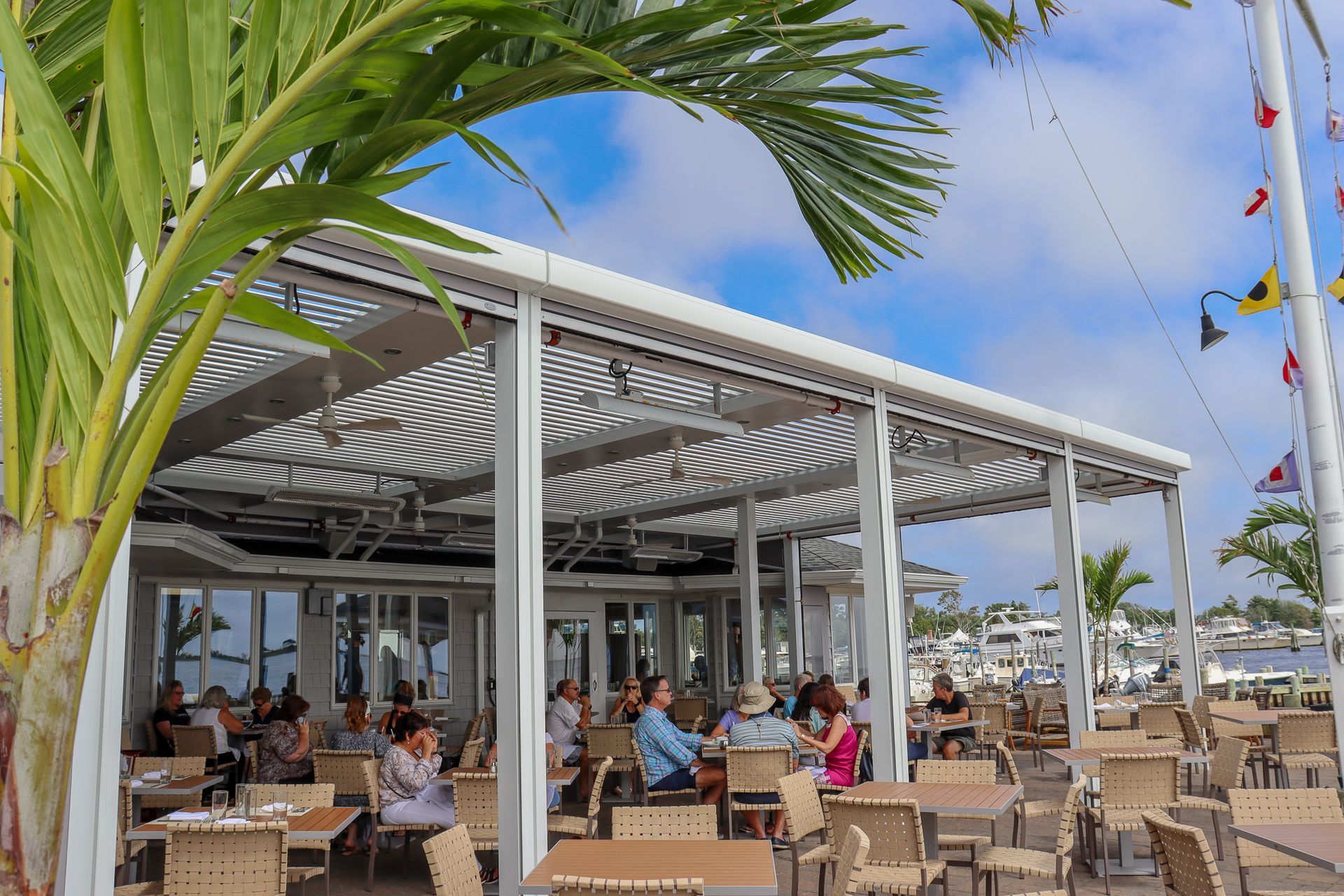 Open-air seaside café with people dining under a striped canopy, palm fronds in the foreground.