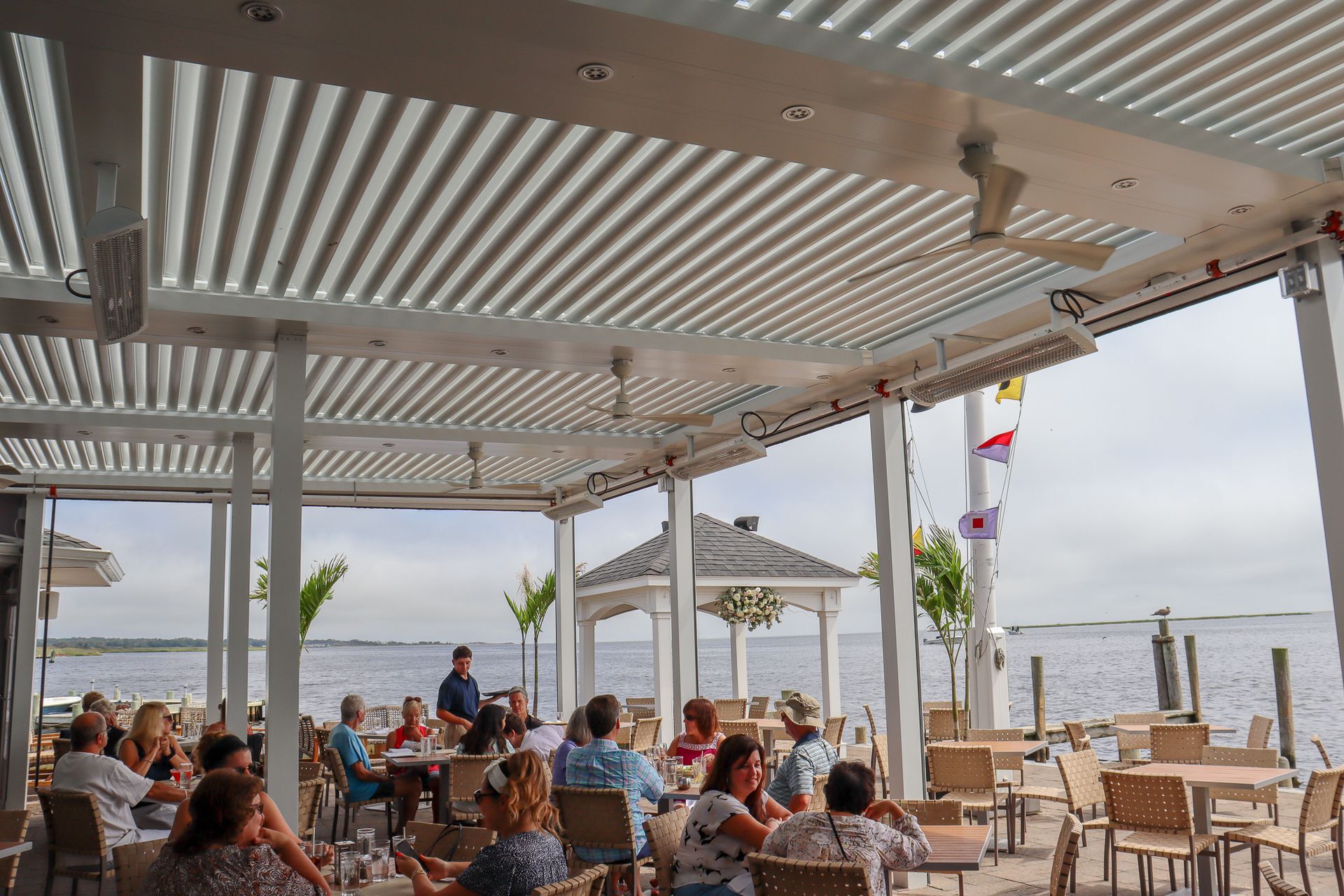 Beachfront café with seated patrons under a striped pergola, overlooking the sea.