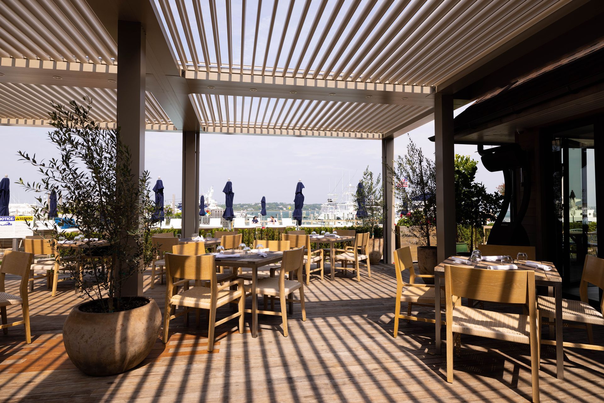 Open-air restaurant patio with wooden tables, chairs, and striped pergola shadows overlooking the water.
