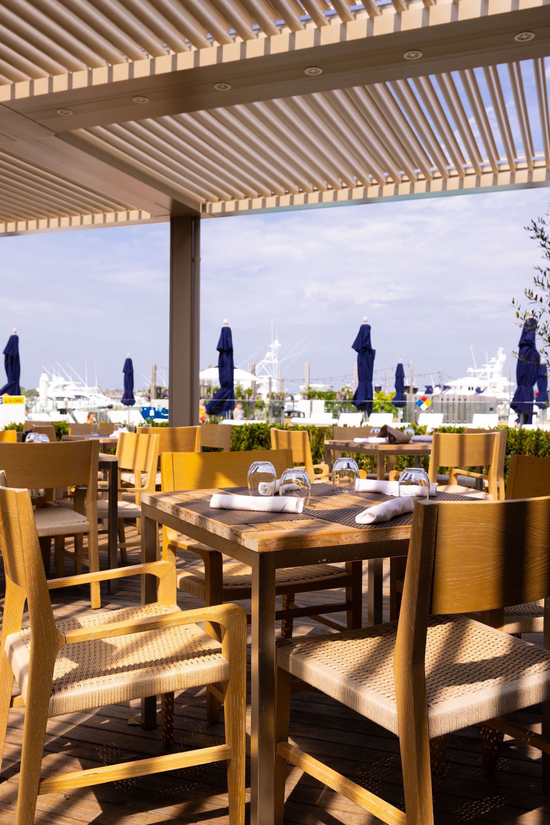 Outdoor seaside restaurant terrace with wicker chairs, tables set for dining, and closed blue umbrellas under a pergola.