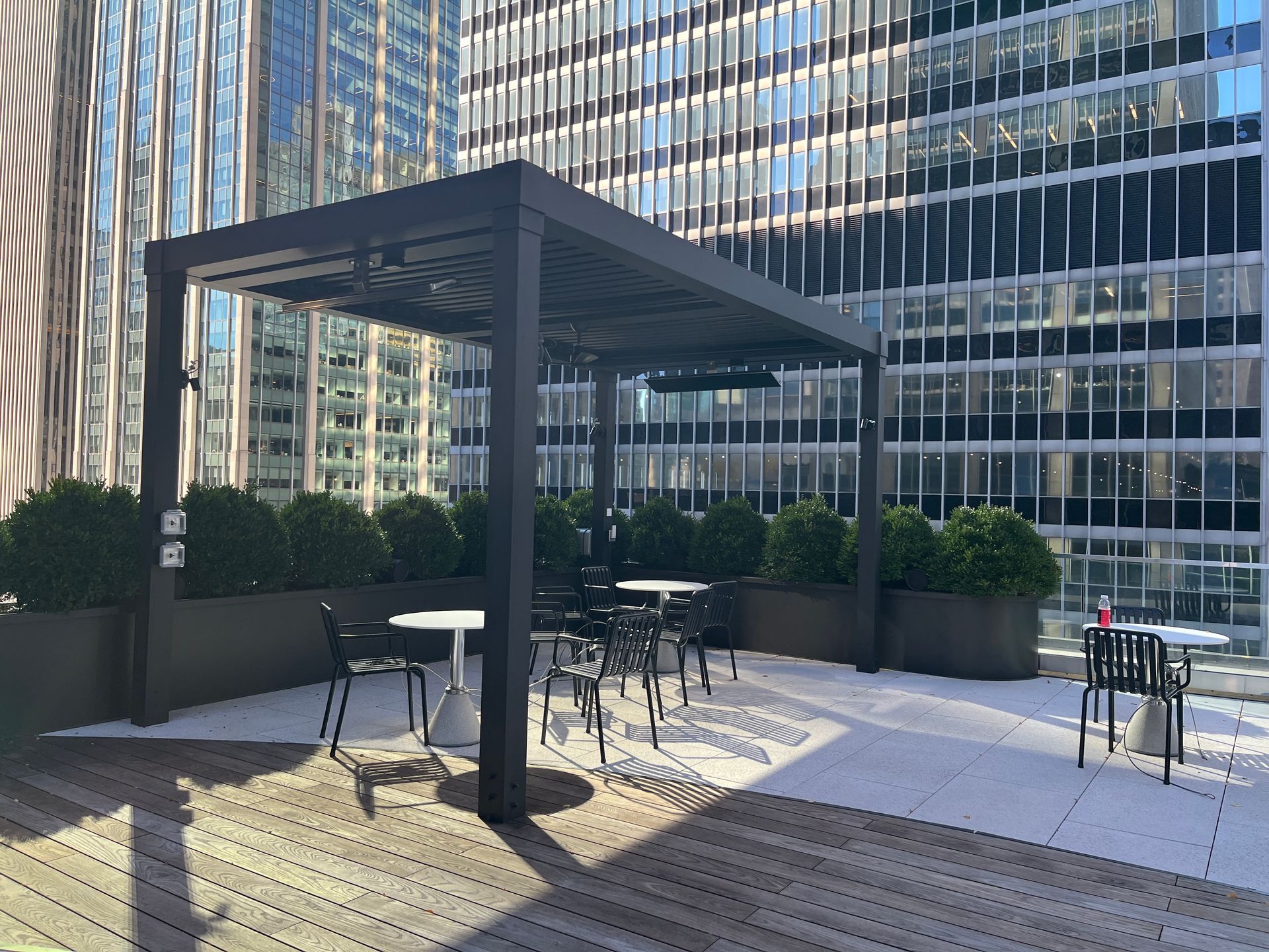 Outdoor café seating under a pergola beside modern glass office buildings.
