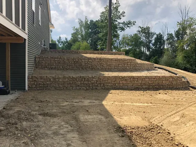 A retaining wall sits on a dirt hill next to a house.