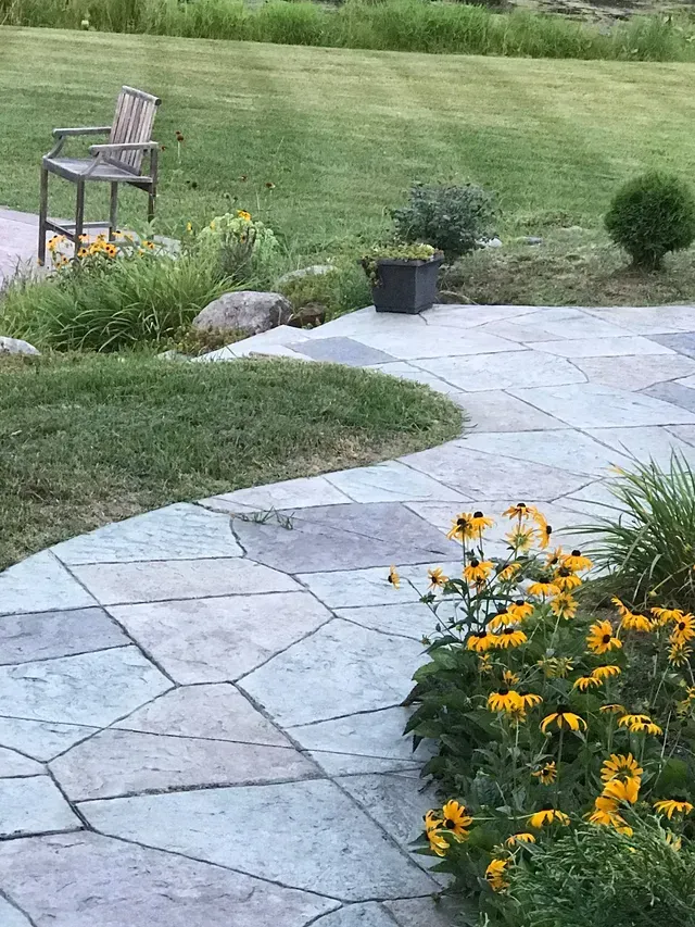 A stone walkway leading to a lawn with flowers and a chair.