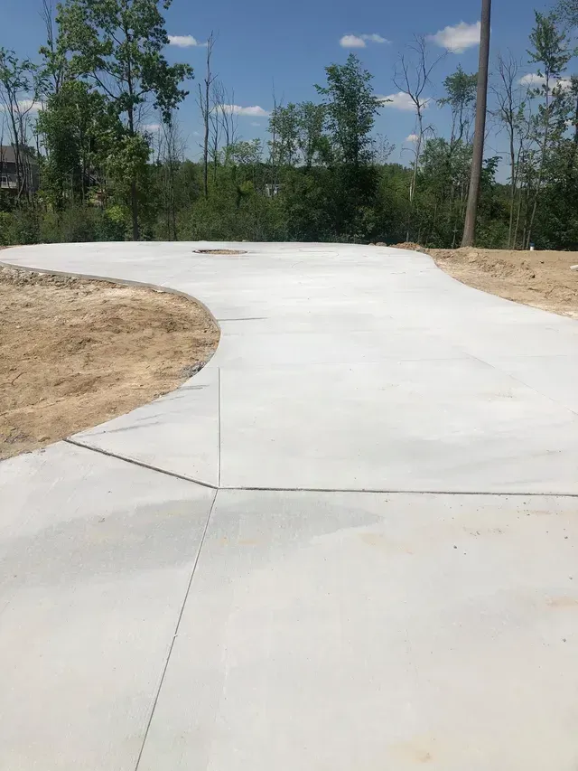 A concrete driveway going through a dirt field with trees in the background.