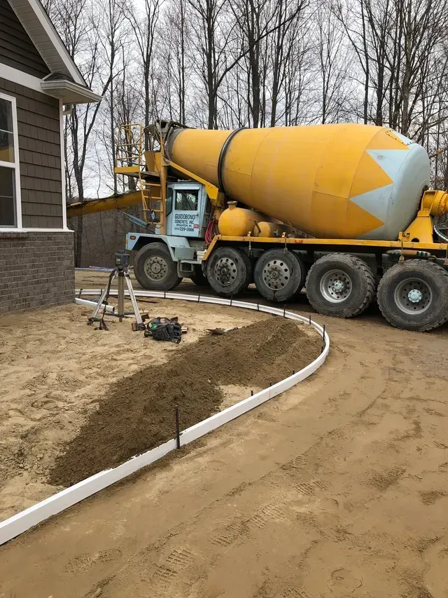 A yellow concrete mixer truck is driving down a dirt road next to a house.