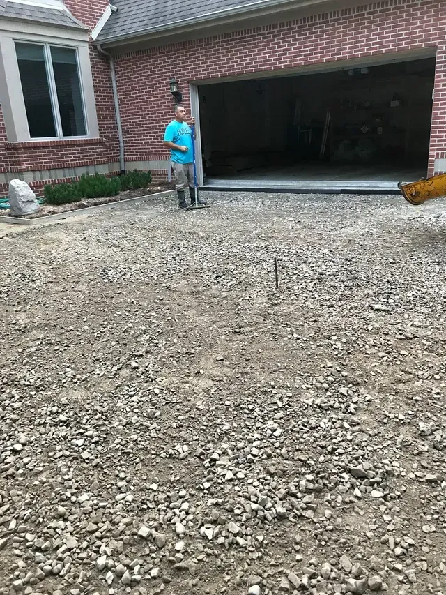 A man is standing in a gravel driveway in front of a brick house.