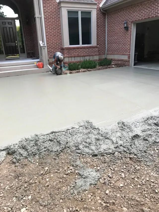 A man is working on a concrete driveway in front of a brick house.